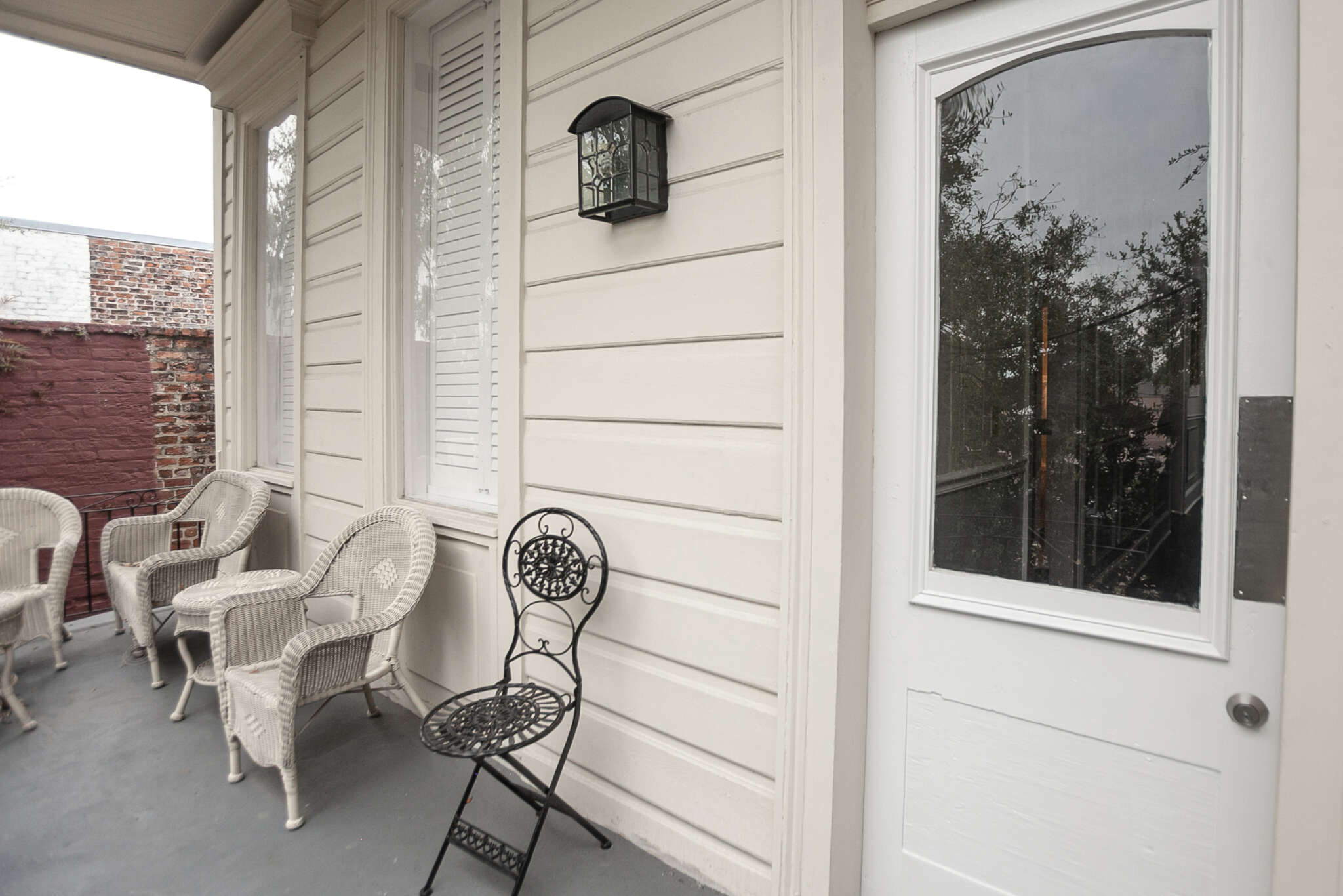 A white porch with wicker chairs and a metal chair, a lantern on the wall, and a door with glass.